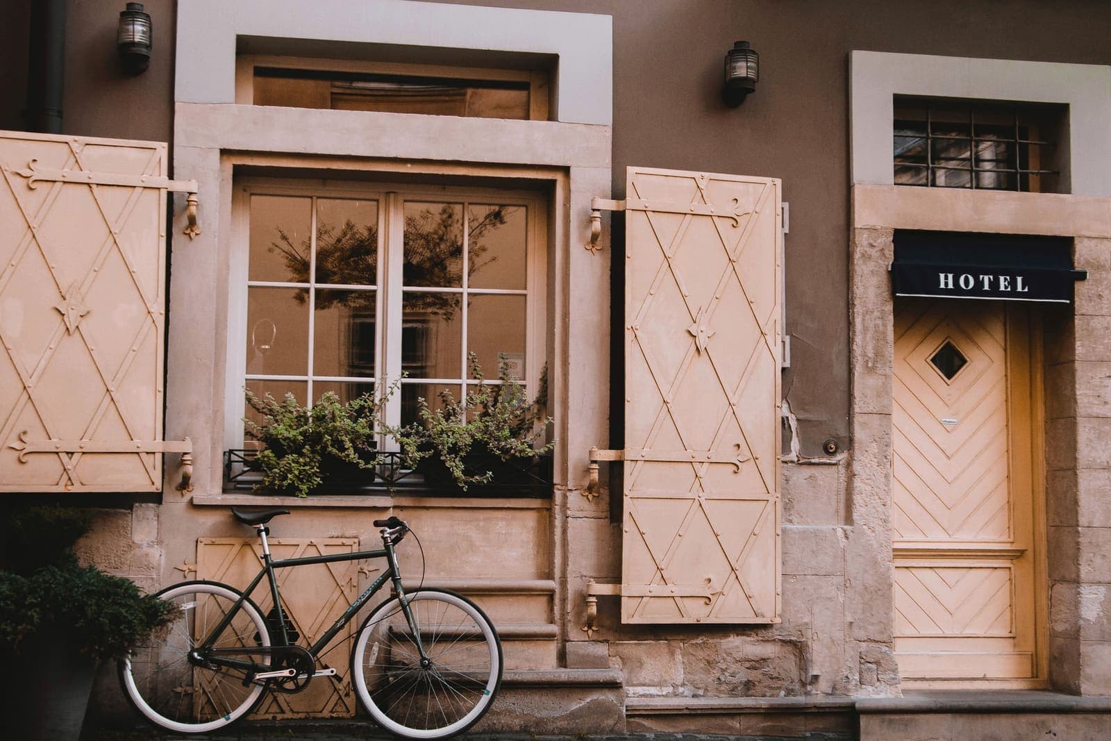 La fachada de un hotel pequeño con contraventanas de madera, flores en la ventana y una bicicleta apoyada en la pared